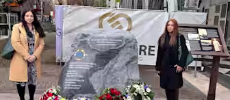 Two women stand on either side of a memorial stone decorated with flowers and wreaths. The stone has an engraved inscription dedicated to those who lost their lives from exposure to asbestos, with a banner and information board in the background.