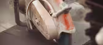 A close-up of a circular saw cutting through a dark sheet of material, with sparks flying from the blade. The scene appears to be part of a construction or fabrication process.
