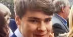 A young man with short, light brown hair is smiling. He is wearing a light blue dress shirt and a dark blazer. The background is slightly blurred, showing a group of people, trees, and some buildings.