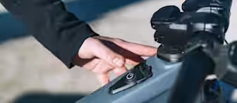 A person's hand, wearing a black sleeve, is pressing the power button on the frame of an electric bicycle near the handlebars.