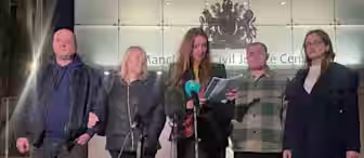 Five people stand in front of the Manchester Civil Justice Centre. A woman in the center holds a speech, with microphones in front of her. The group appears solemn, and it is nighttime. The building's signage and emblem are visible in the background.