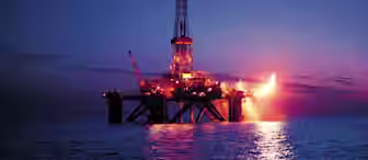 An offshore oil rig stands illuminated against a twilight sky, with flames visibly flaring from one side. The structure is surrounded by calm ocean waters, reflecting the lights from the rig and creating a contrast with the dimming horizon.