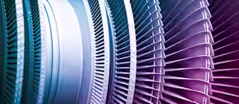 Close-up image of industrial turbine blades, showing a detailed view of the intricate metal components arranged in a circular pattern. The blades display various shades of blue and pink, highlighting the precision and engineering involved in their design.
