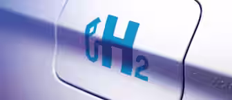Close-up of a car's fuel cap cover with a blue "H₂" symbol, indicating it uses hydrogen as a fuel source. The silver metallic surface of the car is slightly blurred in the background.