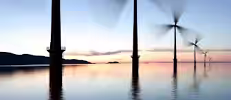 A row of wind turbines standing in calm water during sunset, with blurred blades indicating movement. The sky transitions from light blue to peach and pink near the horizon. Hills are faintly visible in the background.