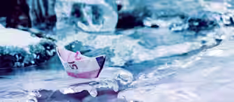 A small paper boat made from a five Euro banknote rests on a piece of ice. The background displays a cold, icy landscape with various ice formations and a frozen water body. The scene highlights the contrast between the colorful banknote and the icy environment.
