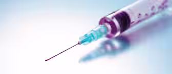 A close-up shot of a syringe with a sharp needle, filled with a red liquid, placed against a smooth gradient background of light blue and white. The syringe is slightly angled, with the needle pointing to the left.