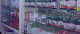 A glass cabinet in a pharmacy displays a variety of bottles and boxes containing vitamins, supplements, and medications. The shelves are neatly organized with products ranging from calcium and vitamin D3 supplements to multivitamins and other health aids.