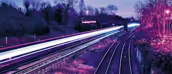 A nighttime scene of a railway with light trails from a passing train. The tracks run through a wooded area with bare trees, and the sky exhibits a purple and blue hue, suggesting dusk or dawn. The long exposure effect captures the movement of the train as bright streaks.