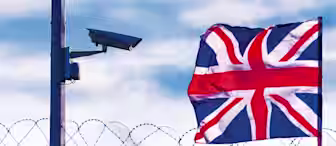 A surveillance camera mounted on a pole overlooks a barbed wire fence. In the background, a Union Jack flag waves against a cloudy sky.