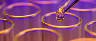 A close-up image shows a pipette dispensing a clear liquid into one of several glass test tubes. The background is blurred with a mix of blue and purple hues, creating a scientific and vibrant atmosphere.