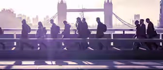 Silhouettes of people walking across a bridge during early morning with the iconic Tower Bridge in London visible in the background. The sky is filled with a soft glow from the rising sun.