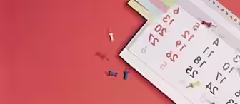 A desk calendar turned to the month of March, with several colored sticky notes attached to the top. Red, blue, yellow, and white thumbtacks are scattered on the red surface around the calendar.