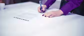 A person wearing a purple shirt is signing a contract on a white table with a pen. Another person in a blue suit stands opposite, with only their torso and hands visible. A small white coffee cup is placed on the table in the background.