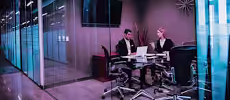 Two people in business attire sit at a conference table in a modern glass-walled office, working on a laptop and discussing documents. The room features a TV, modern decor, and office chairs.