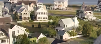 A coastal neighborhood with several large, light-colored houses near a body of water. The homes have various architectural designs, with porches and large windows, surrounded by greenery and white fences. A boat is seen in the water in the distance.