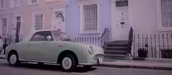 A pastel-colored neighborhood street features a vintage, mint green car parked in front of a row of charming terraced houses. The houses have pastel-colored facades, white trims, and black iron railings, creating a picturesque and nostalgic scene.