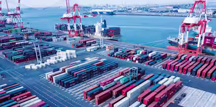 Aerial view of a busy shipping port with colorful stacked shipping containers, large cranes, and docked cargo ships. The blue water of the bay and distant industrial cityscape are visible in the background.