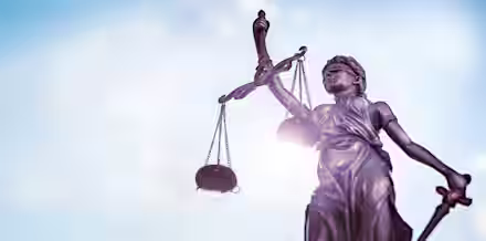 A bronze statue of Lady Justice holding scales in her left hand and a sword in her right hand, with a blindfold covering her eyes. The background is a clear, bright blue sky with light clouds. The image focuses on the statue, symbolizing fairness and law.