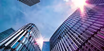 A low-angle view of modern office buildings with reflective glass and metal facades, rising towards a bright sky. The sun is shining brightly, creating lens flare effects on the buildings. The architectural structures are sleek and contemporary.