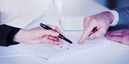 Two people sit at a table reviewing a document with charts and graphs. One person is holding a pen and gesturing towards the paper, while the other person points with their finger. A laptop and a glass of water are visible in the blurred background.