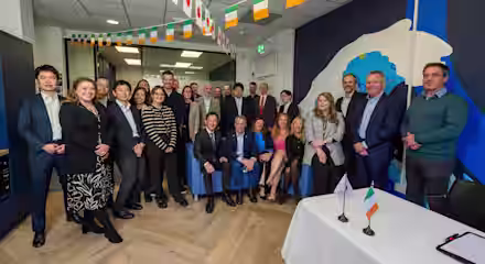 A group of people poses for a photo in an office decorated with Japanese and Irish flags; two small flags are placed on a white table in the foreground.