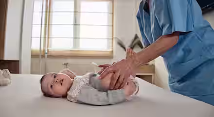A baby lies on an examination table while an adult in blue scrubs gently holds and moves the baby’s legs, likely during a medical or physical therapy checkup in a bright room.