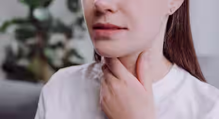 A woman in a white shirt touches her throat with one hand, suggesting discomfort or a sore throat. The background is blurred, with a green plant visible.