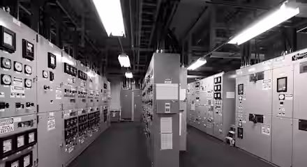 A monochrome image of an industrial control room. The room is filled with large electrical panels, meters, switches, and labels. Overhead fluorescent lights illuminate the space, casting shadows on the floor.