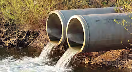 Two large concrete pipes discharge water into a natural stream, surrounded by greenery and branches. The emerging water creates ripples and bubbles on the stream's surface, indicating the flow from the pipes.