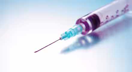 A close-up shot of a syringe with a sharp needle, filled with a red liquid, placed against a smooth gradient background of light blue and white. The syringe is slightly angled, with the needle pointing to the left.