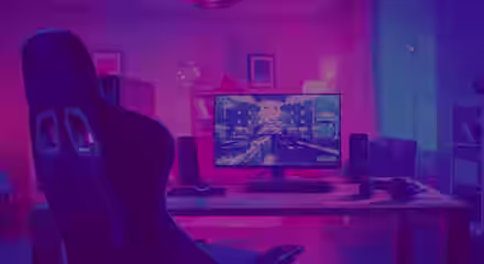 A gaming setup in a dimly lit room featuring a desk with a large monitor, keyboard, and mouse. The screen displays a futuristic game. Surround sound speakers, headphones, and a gaming chair are also visible, all bathed in pink and blue ambient lighting.