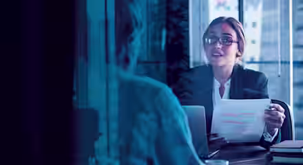 A woman wearing glasses and a business suit is holding a paper and speaking to another person across a desk in an office setting. The scene is viewed through a window, reflecting the cityscape outside.