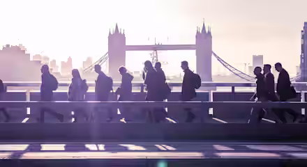 Silhouettes of people walking across a bridge during early morning with the iconic Tower Bridge in London visible in the background. The sky is filled with a soft glow from the rising sun.
