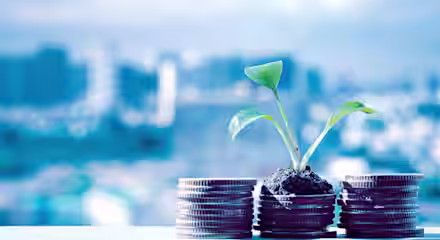 A small plant grows out of a pile of soil on top of a stack of coins, set against a blurred cityscape background. The scene symbolizes financial growth and investment in sustainability.