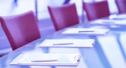A glass conference table is set with clipboards, pens, and papers. Red chairs are arranged around the table in the background, with a soft focus on the modern, light-filled space. The overall tone of the image is cool, with blue and purple hues.