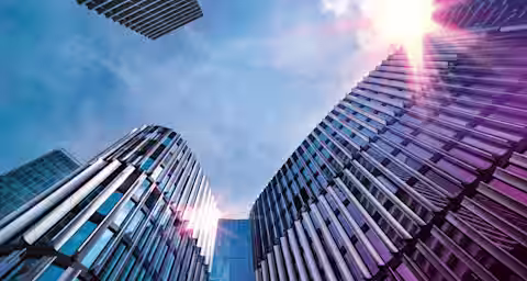 A low-angle view of modern office buildings with reflective glass and metal facades, rising towards a bright sky. The sun is shining brightly, creating lens flare effects on the buildings. The architectural structures are sleek and contemporary.