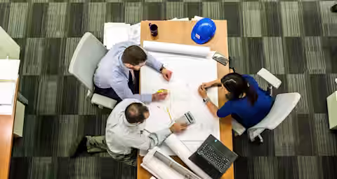 Three people are seated around a table with large architectural blueprints. One person is using a calculator, another is writing with a pencil, and the third has a laptop open. A blue hard hat and a smartphone are also on the table. The room has a striped carpet.