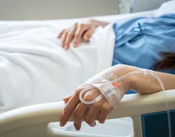 A person lies in a hospital bed, covered with a white blanket. Their hand, with an intravenous (IV) tube inserted and secured by transparent tape, rests on the bed railing. They are wearing a blue hospital gown, partially visible in the image.