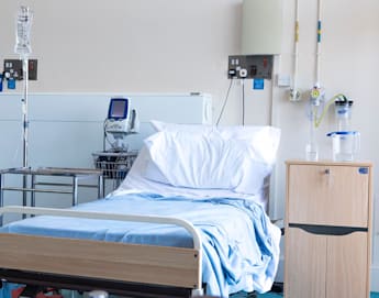 A hospital room with a single bed, white pillows, and light blue sheets. An IV stand and medical equipment are positioned next to the bed. A water jug and glass rest on a small wooden bedside table. The room appears clean and orderly.