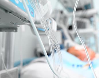 A hospital room with a blurred patient lying in bed in the background. Various medical equipment and monitoring devices, including IV lines and tubes, are focused in the foreground. The scene suggests a critical care or intensive care situation.