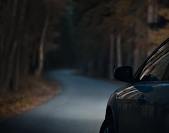 A dark road curves through a forest with tall trees, illuminated faintly by a distant light. The silhouette of a car is visible on the right side of the image, suggesting it is parked or driving along the road. The scene is moody and atmospheric.