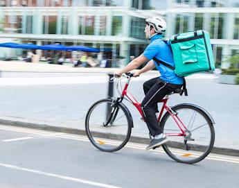 A delivery person rides a red bicycle on a city street, wearing a blue shirt, black pants, a white helmet, and carrying a large green insulated backpack. Blurred buildings and outdoor seating areas are visible in the background.