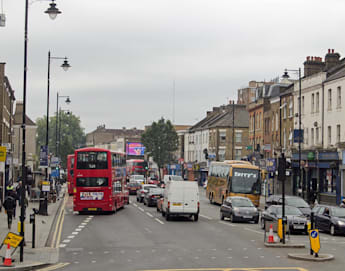 A busy city street with a red double-decker bus, a tour bus, several cars, and vans in traffic. Pedestrians are walking on the sidewalks lined with buildings and shops. Street lights and signs are visible, and the sky is overcast.