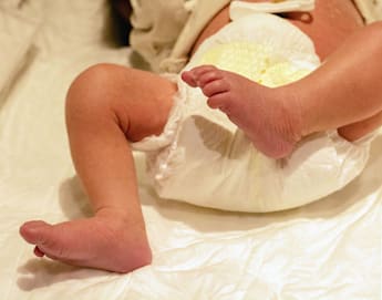 A newborn baby wearing a yellow diaper rests on a white blanket. The baby's legs are slightly bent, with the left foot lifted higher than the right. The focus is on the lower half of the baby's body, emphasizing the delicate and tiny feet.