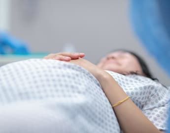 Close-up of a pregnant woman lying on a hospital bed, wearing a patterned hospital gown. Her hands are gently resting on her belly. The background shows medical staff wearing blue gloves.