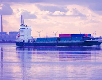 A cargo ship carrying stacked shipping containers sails on calm waters near an industrial cityscape. Wind turbines and factory buildings are visible in the background under a cloudy sky with a purple-pink tint.