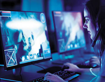 A person wearing a headset sits at a computer, engaged in a video game. The room is dimly lit with a blue hue. Multiple monitors display gameplay, featuring vibrant graphics. The person uses a keyboard and mouse, focused on the screen.