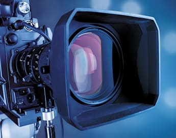 Close-up shot of a professional video camera lens with a large square hood, capturing a clear view of the lens glass. The background is blurred and has a gradient of blue tones with lights creating bokeh effects.