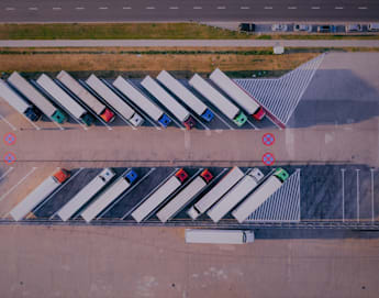 Aerial view of a parking lot with several semi-trucks parked diagonally in designated spaces. A single truck is parked perpendicularly at the lot's edge. There are red "X" markings indicating no-parking zones, and a road with moving vehicles is visible nearby.